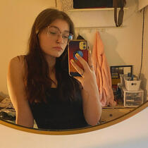 A woman with long brunette hair and glasses taking a selfie in a mirror. She's wearing a black tank top and she's looking at her phone screen.